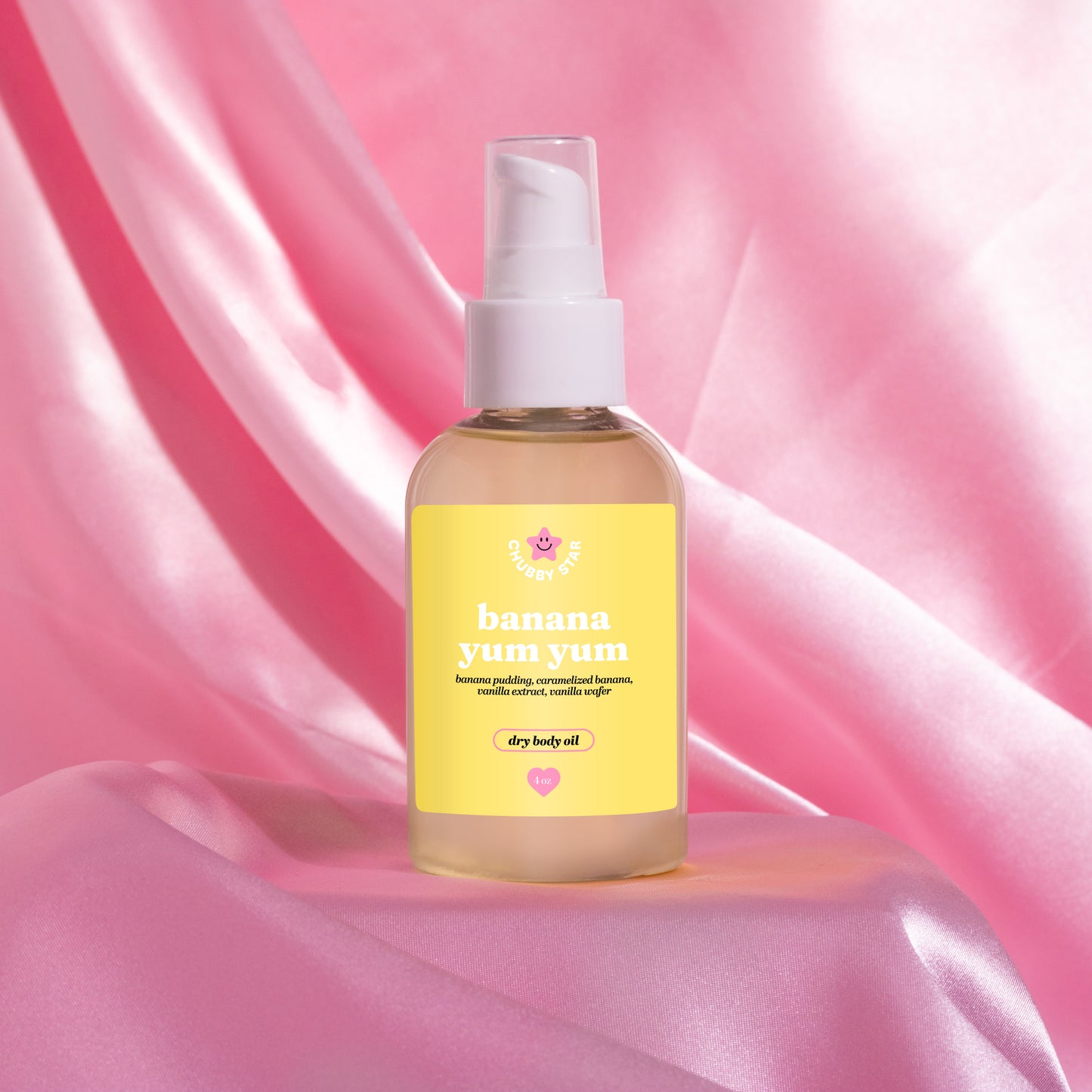 A bottle of banana yum dry body oil with a yellow label and white pump dispenser, set against a draped pink satin fabric background.