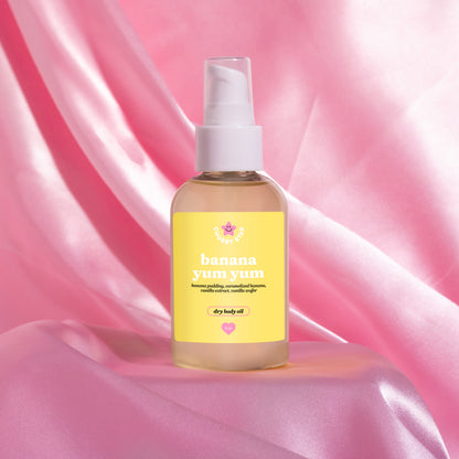 A bottle of banana yum dry body oil with a yellow label and white pump dispenser, set against a draped pink satin fabric background.