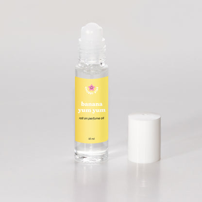 A 10 ml clear glass rollerball bottle of banana yum yum roll-on perfume oil with a yellow label, shown next to its white cap on a light gray background.