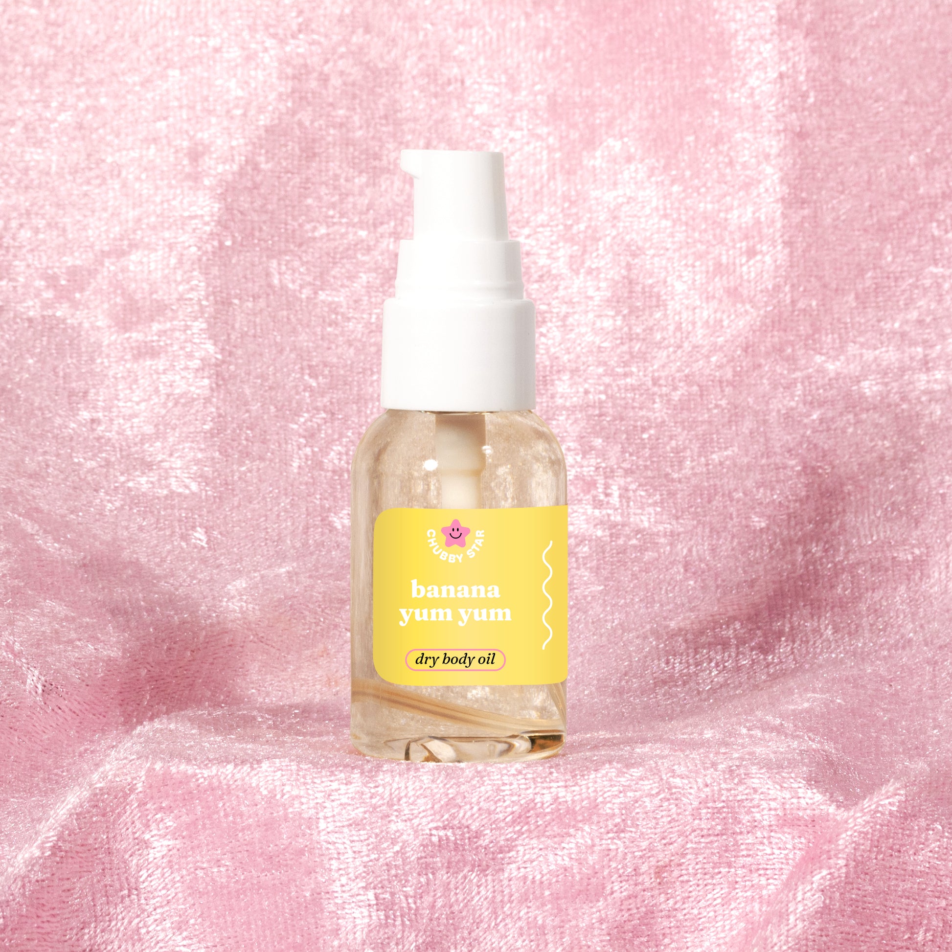 A clear mini pump bottle of 'banana yum yum' body oil by the brand 'chubby star' with a white pump and a yellow colored label, set against a textured pink fabric background.