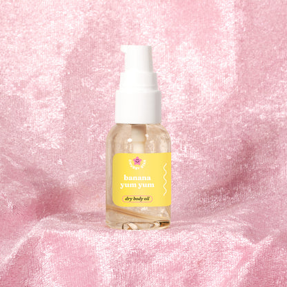 A clear mini pump bottle of 'banana yum yum' body oil by the brand 'chubby star' with a white pump and a yellow colored label, set against a textured pink fabric background.
