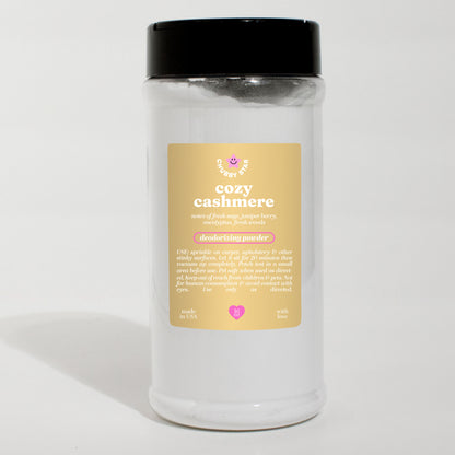 A white clear jar of 'cozy cashmere' scented carpet freshening deodorizing powder with a black lid and a beige label, set against a plain white background.