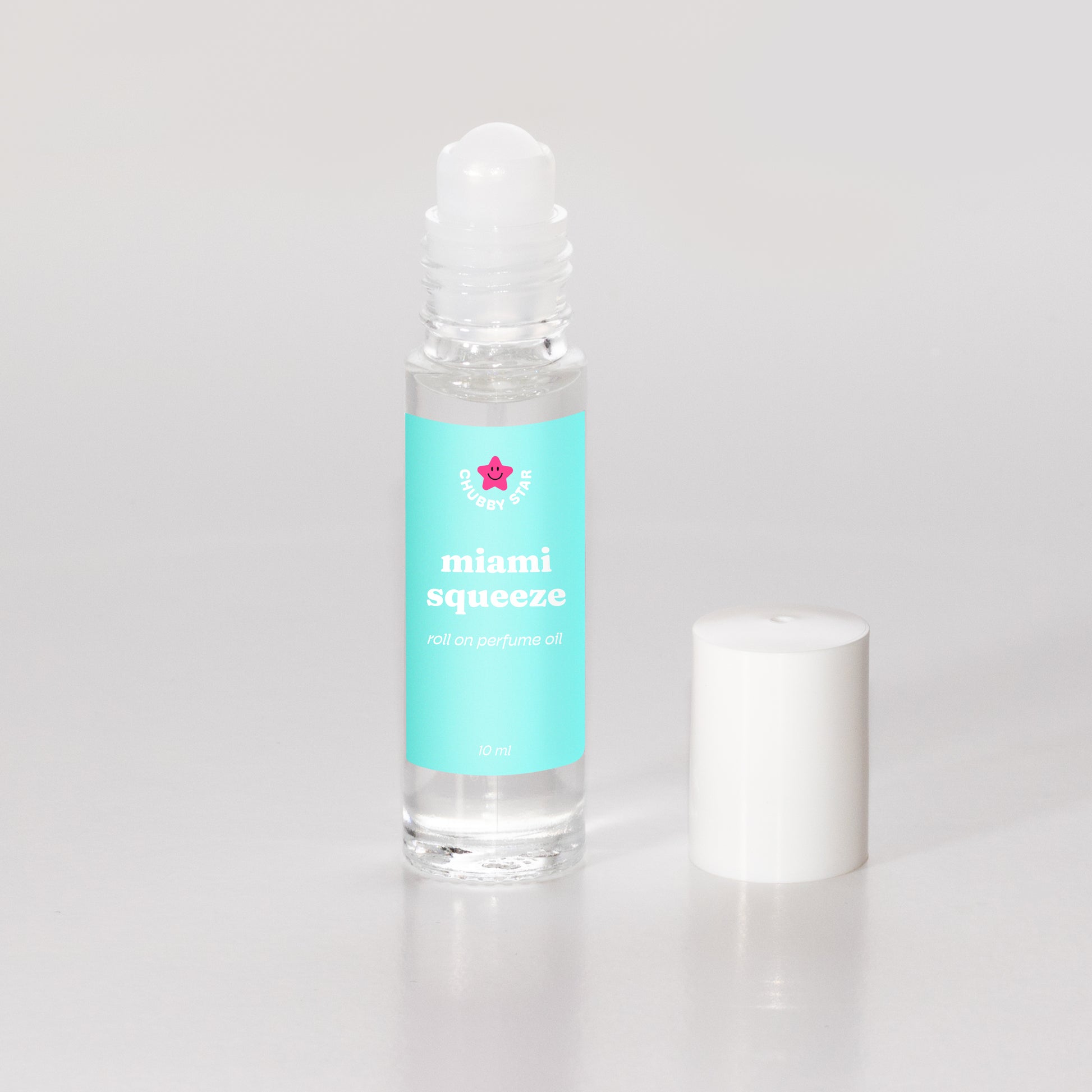 A clear glass rollerball perfume oil bottle filled with transparent oil, featuring a turquoise label with the scent 'miami squeeze aka miami nectar' and a small pink star logo, shown next to its white cap on a light gray background