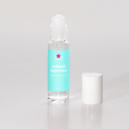 A clear glass rollerball perfume oil bottle filled with transparent oil, featuring a turquoise label with the scent 'miami squeeze aka miami nectar' and a small pink star logo, shown next to its white cap on a light gray background