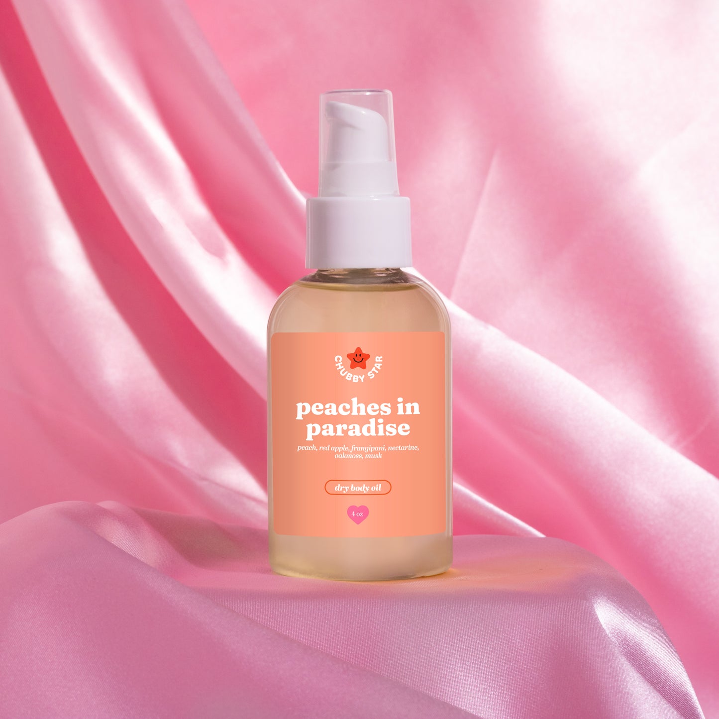 A bottle of body oil with a white pump dispenser and a peach-colored label reading 'peaches in paradise' inspired by the kayali eden sweet peach 35 scent, set against a draped pink satin background.