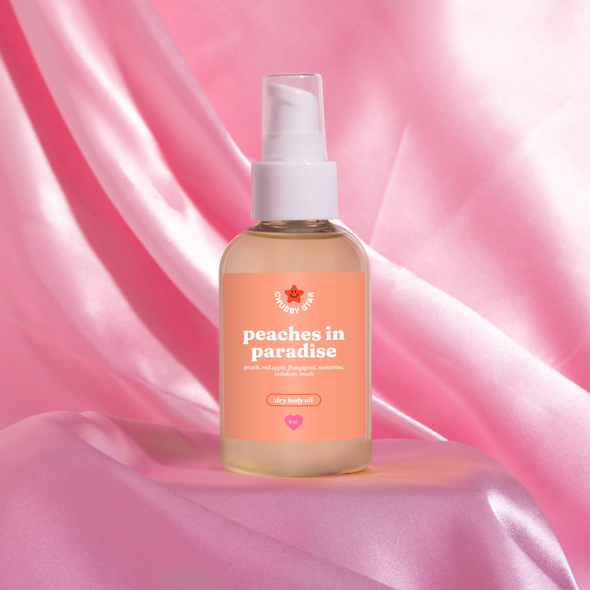A bottle of body oil with a white pump dispenser and a peach-colored label reading 'peaches in paradise' inspired by the kayali eden sweet peach 35 scent, set against a draped pink satin background.