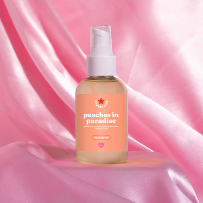 A bottle of body oil with a white pump dispenser and a peach-colored label reading 'peaches in paradise' inspired by the kayali eden sweet peach 35 scent, set against a draped pink satin background.