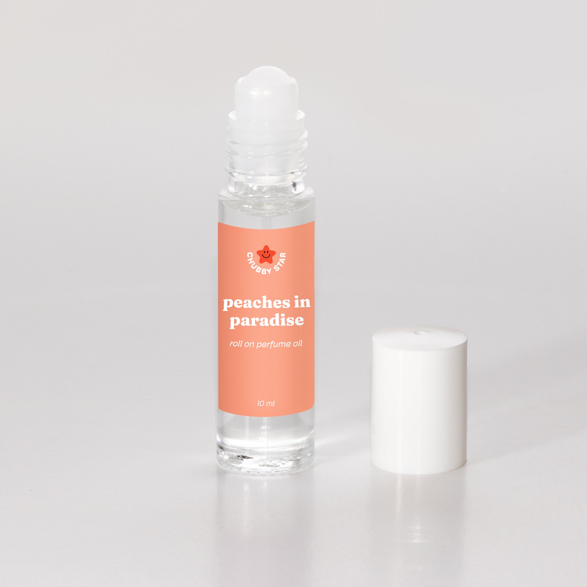 A clear glass rollerball perfume oil bottle filled with transparent oil, featuring a peach colored label with the scent 'peaches in paradise, inspired by kayali eden sweet peach' and a small star logo, shown next to its white cap on a light gray background