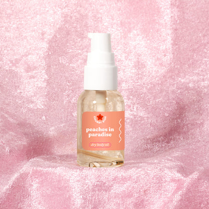 A clear mini pump bottle of 'peaches in paradise' body oil inspired by kayali eden sweet peach 35 by the brand 'chubby star' with a white pump and a peach-colored label, set against a textured pink fabric background.